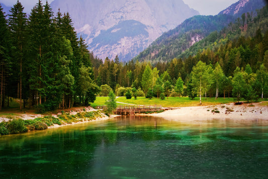 Scenic landscape print of a jasna lake with clear green and blue water, a sandy shore, a small wooden bridge, dense evergreen trees, and rocky mountains in the background.