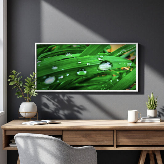 A framed wall art print depicting vibrant green leaves with water droplets, displayed on a dark gray wall above a wooden desk with plants and a mug.