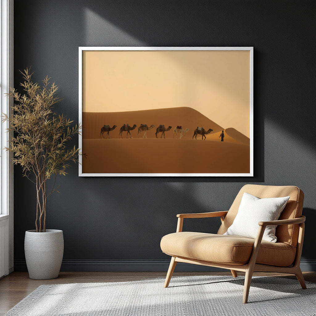 A framed desert art print hangs on a dark charcoal wall above a tan armchair with a white pillow, next to a potted plant. The print shows a caravan of camels on golden sand dunes under a warm beige sky.