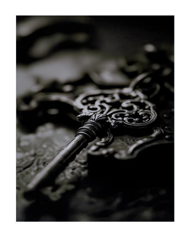 A close-up photograph of an ornate antique-style metal key resting on a dark, textured surface with shallow depth of field.