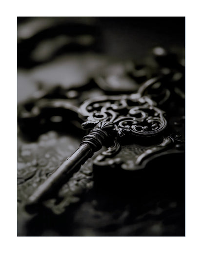 A close-up photograph of an ornate antique-style metal key resting on a dark, textured surface with shallow depth of field.