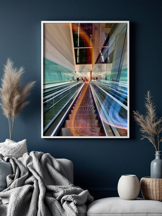 A framed abstract art print depicting escalators with colorful light trails, displayed on a dark blue wall above a modern gray sofa with a textured throw blanket and decorative dried grass in vases.