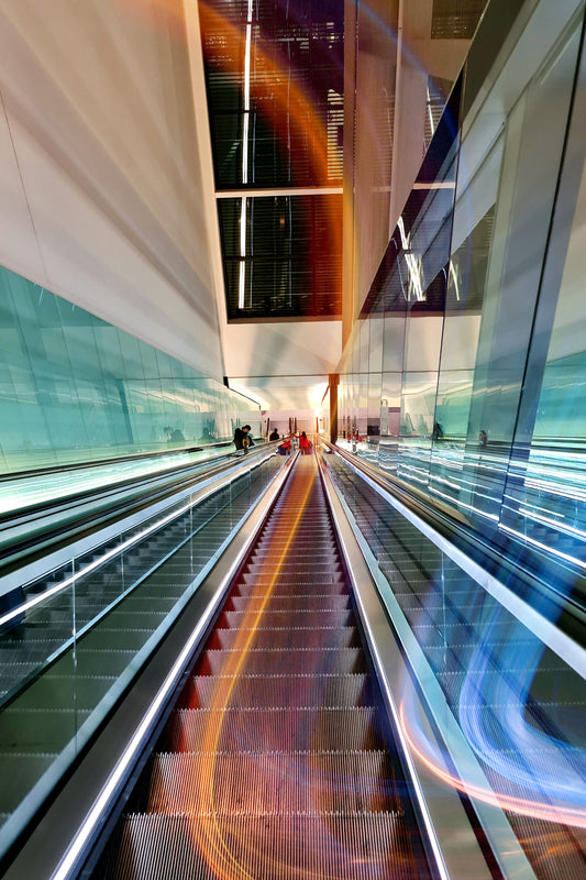 A long-exposure photograph of an escalator in a modern indoor transit or shopping center, with bright light trails in orange, blue, and white reflecting off the glass walls and metal rails.