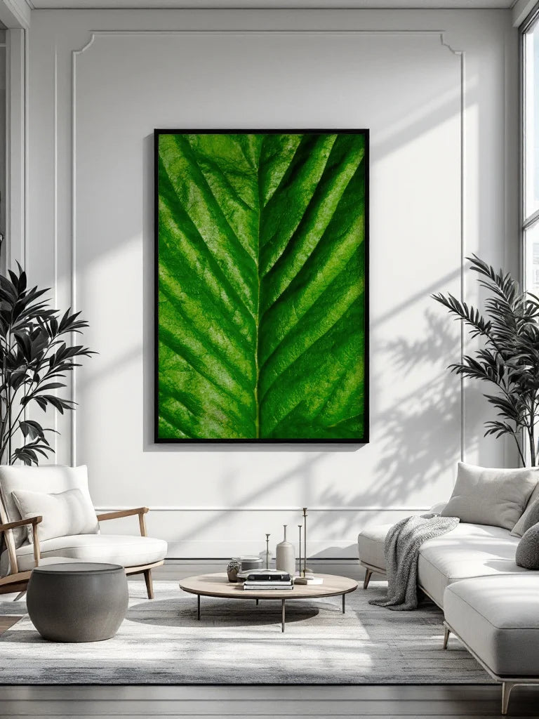 A large framed wall art print featuring a close-up green leaf pattern hangs on a white paneled wall in a modern living room with a sofa, chair, coffee table, and plants.