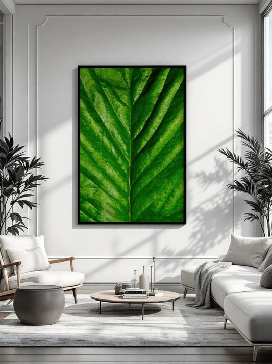 A large framed wall art print featuring a close-up green leaf pattern hangs on a white paneled wall in a modern living room with a sofa, chair, coffee table, and plants.