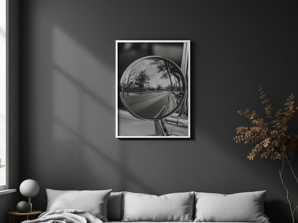 A framed black and white art print depicting a car side-view mirror reflecting a road and trees, displayed on a dark gray wall above a modern sofa in a styled living room.