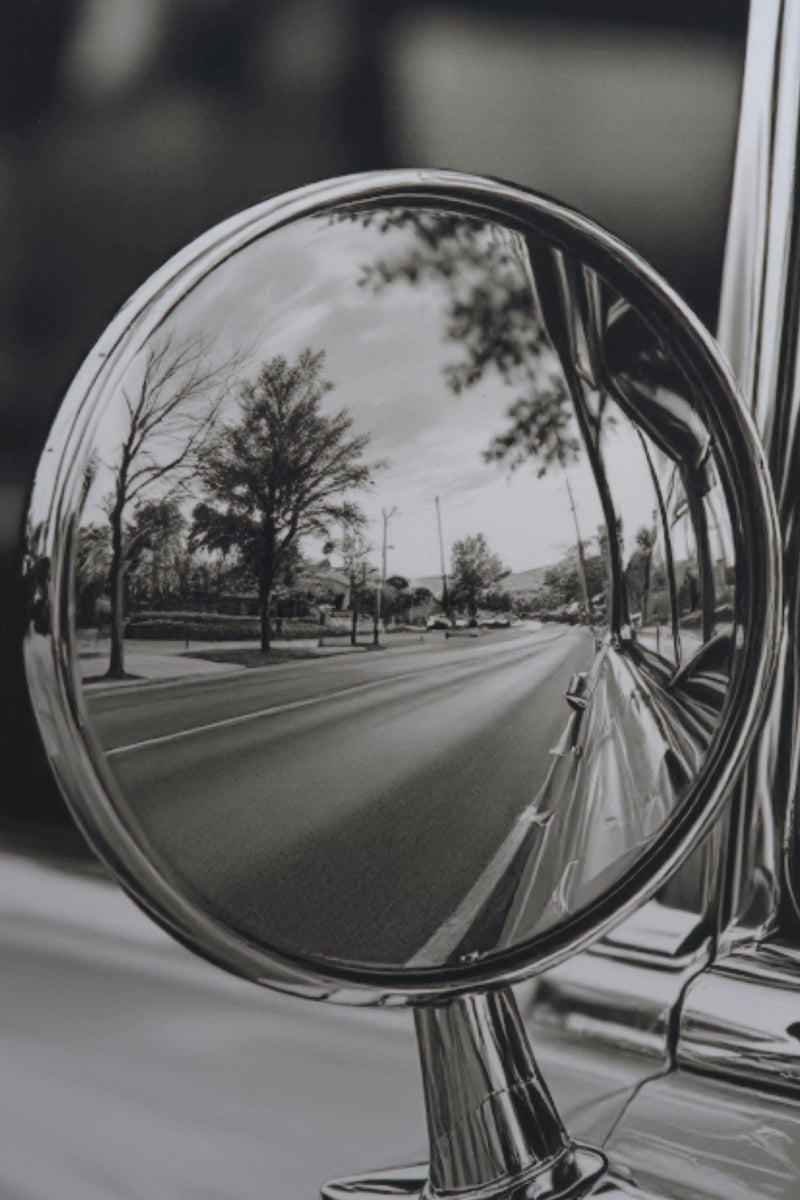 A close-up black and white photograph of a round chrome side-view mirror attached to a car, reflecting a tree-lined road.