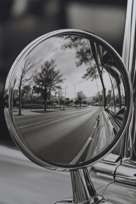 A close-up black and white photograph of a round chrome side-view mirror attached to a car, reflecting a tree-lined road.