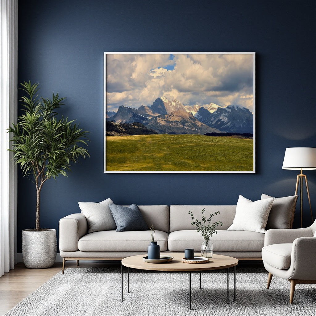 A large framed landscape art print of snow-capped mountains over a green valley under a cloudy sky, displayed on a dark blue wall above a modern light gray sofa in a styled living room.