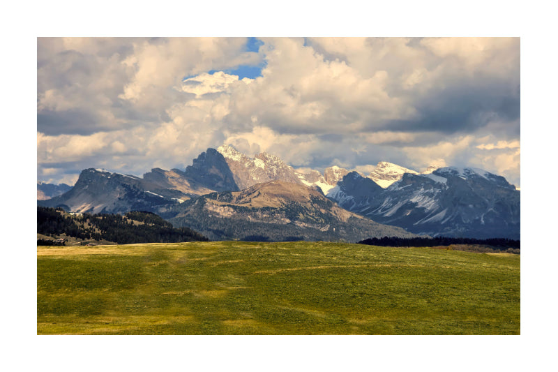 A scenic landscape photograph of rugged snow-capped mountains under a dramatic cloudy sky, with a green valley in the foreground, shown as a square print centered on a white background.