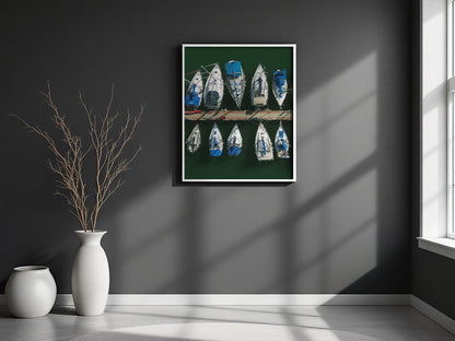 A framed wall art print showing a row of blue and white boats on a dark green background, displayed on a dark gray wall in a modern room setting with a window, vase, and dried branches.