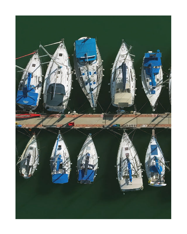 Aerial view of multiple white sailboats with blue covers docked side by side along a pier over dark green water.