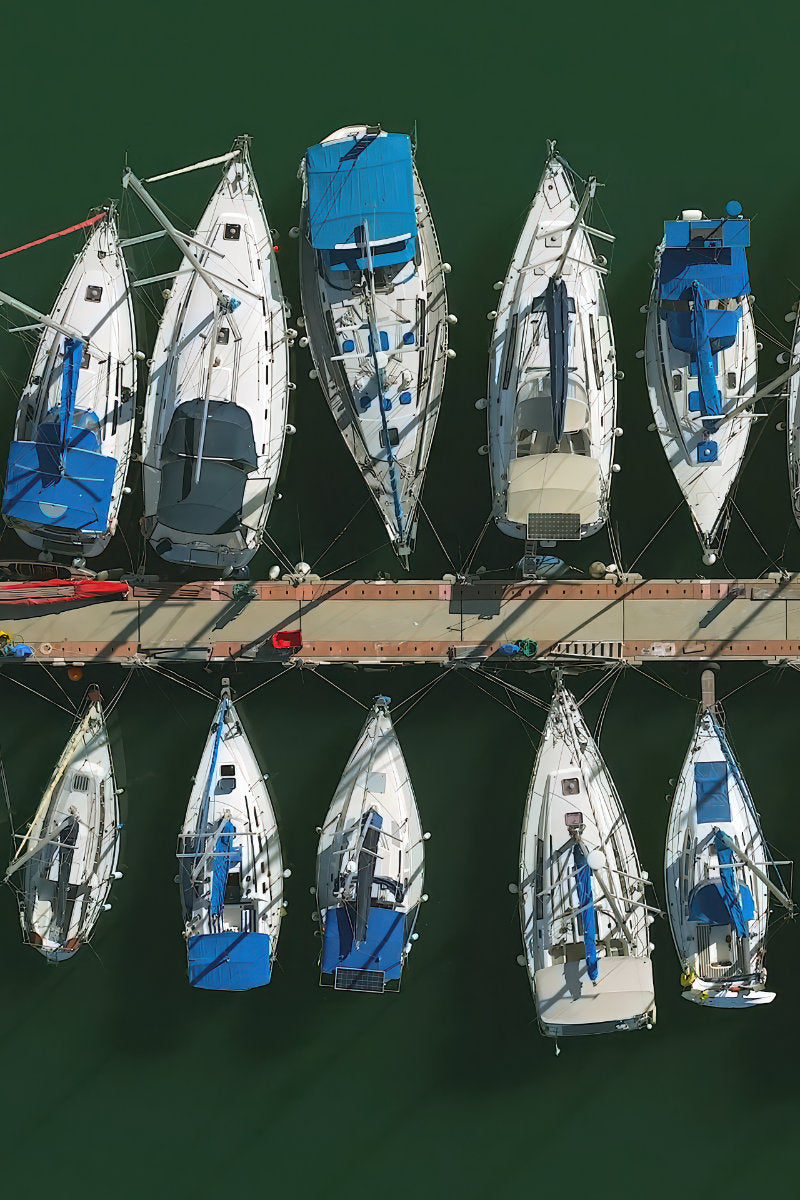 Aerial overhead view of multiple white sailboats with blue covers and rigging moored in a marina on dark green water.