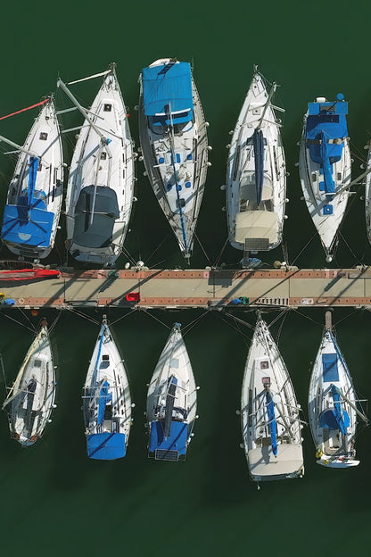 Aerial overhead view of multiple white sailboats with blue covers and rigging moored in a marina on dark green water.