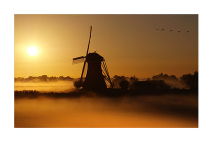 A scenic landscape print featuring a dark silhouette of a windmill standing in misty lowland fog at sunrise, with a bright yellow sun on the left and birds in the sky above, framed by a white border.