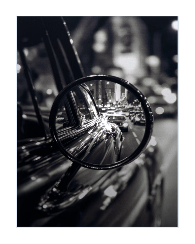 Black and white photographic print featuring a close-up of a classic car side mirror reflecting a blurred city street at night.