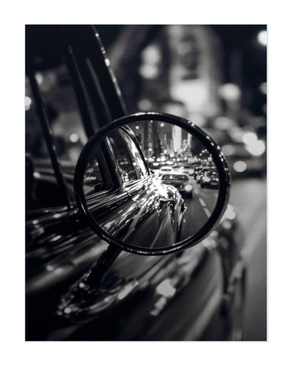 Black and white photographic print featuring a close-up of a classic car side mirror reflecting a blurred city street at night.