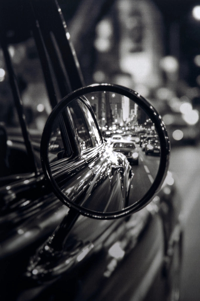 Black and white close-up photograph of a car side-view mirror reflecting a busy city street at night.