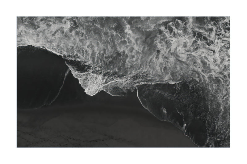 A black and white aerial photograph of ocean waves crashing onto a dark beach, presented as a square print with a white border.