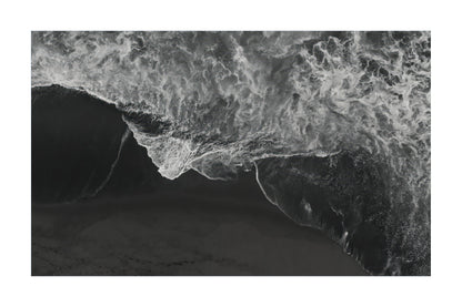A black and white aerial photograph of ocean waves crashing onto a dark beach, presented as a square print with a white border.