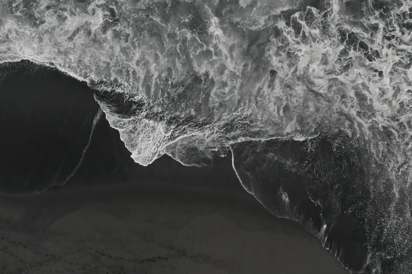 Black and white aerial photograph of a dramatic ocean wave curling over dark sand, with high-contrast white foam and deep gray-black water.