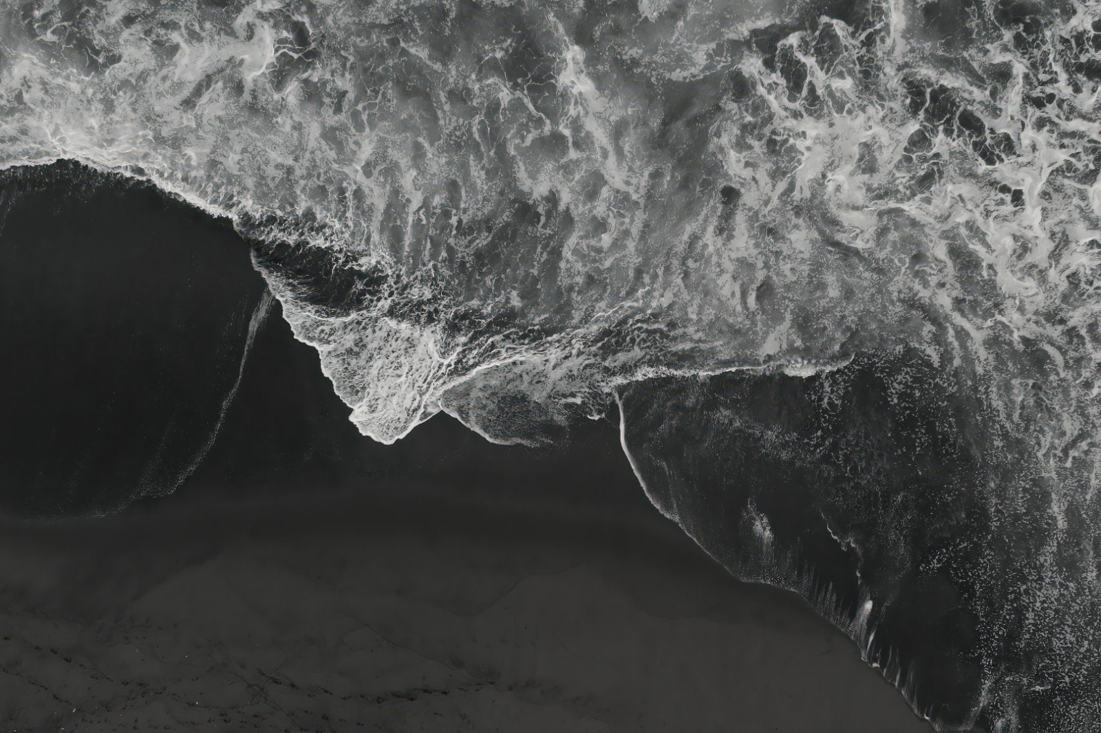 Black and white aerial photograph of a dramatic ocean wave curling over dark sand, with high-contrast white foam and deep gray-black water.