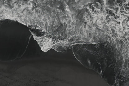 Black and white aerial photograph of a dramatic ocean wave curling over dark sand, with high-contrast white foam and deep gray-black water.