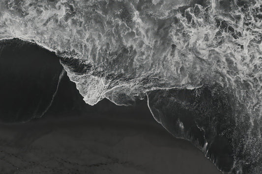 Black and white aerial photograph of a dramatic ocean wave curling over dark sand, with high-contrast white foam and deep gray-black water.