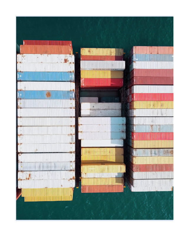 Overhead view of stacked shipping containers in white, blue, yellow, red, and gray on a cargo ship, surrounded by deep teal water.