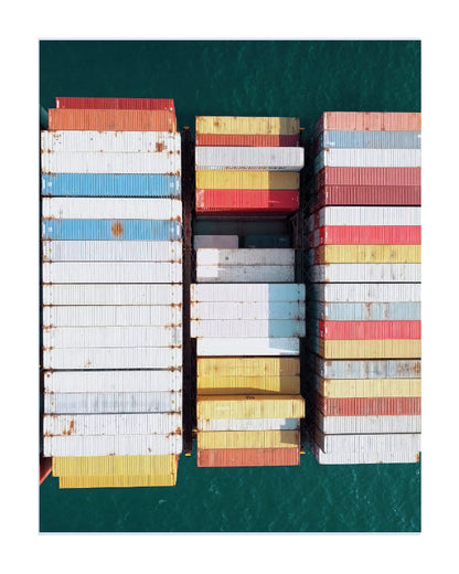 Overhead view of stacked shipping containers in white, blue, yellow, red, and gray on a cargo ship, surrounded by deep teal water.