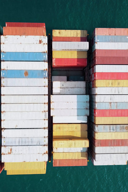 Aerial view of stacked shipping containers in three columns on a cargo ship, surrounded by deep teal water.