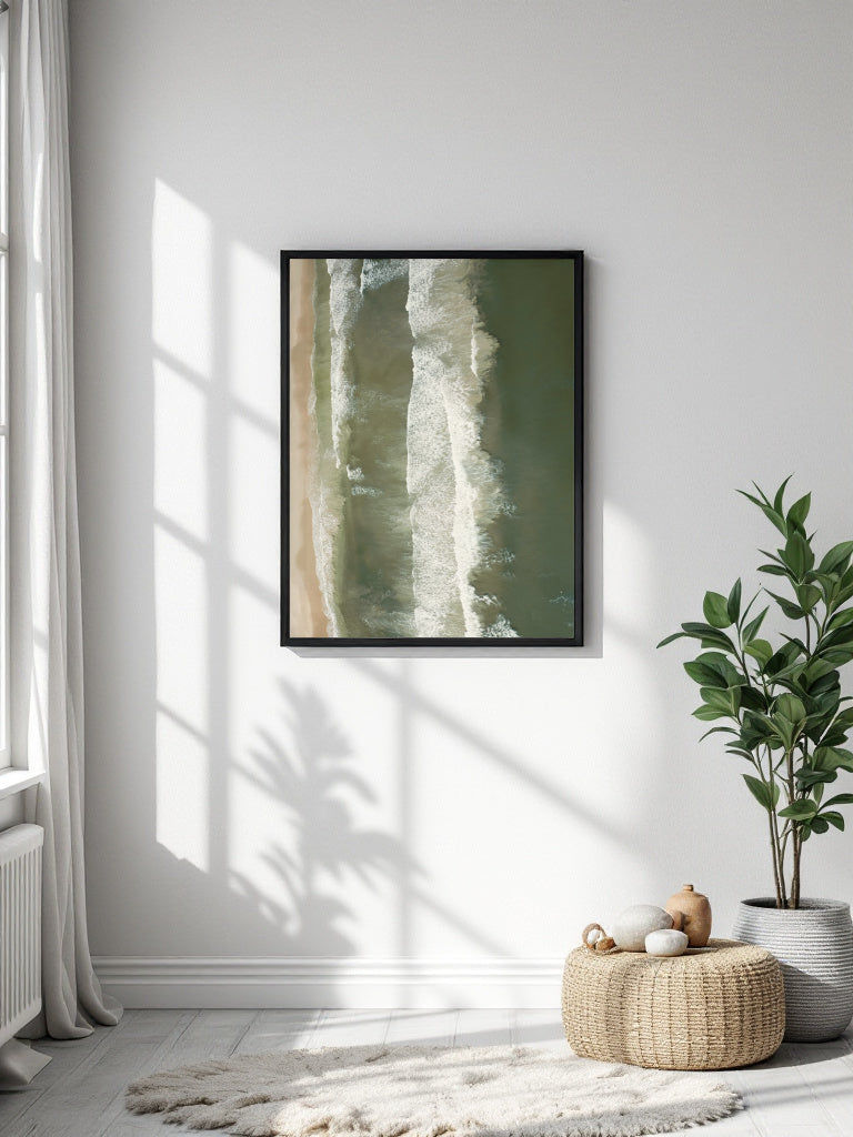 A framed abstract art print with vertical olive green and cream aerial photograph of ocean waves rolling onto a sandy beach hanging on a white wall in a bright room with a potted plant, woven pouf, and soft shadows.