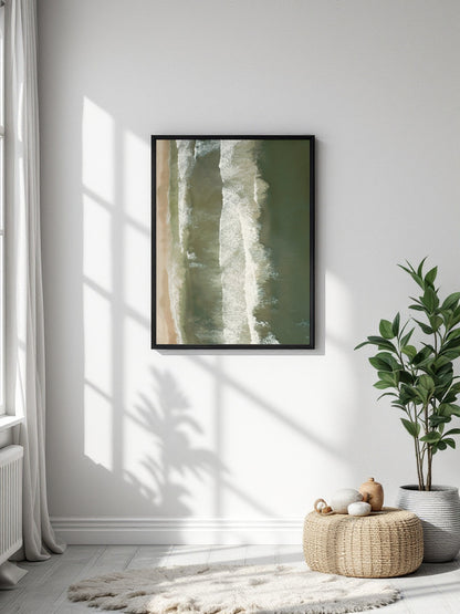 A framed abstract art print with vertical olive green and cream aerial photograph of ocean waves rolling onto a sandy beach hanging on a white wall in a bright room with a potted plant, woven pouf, and soft shadows.