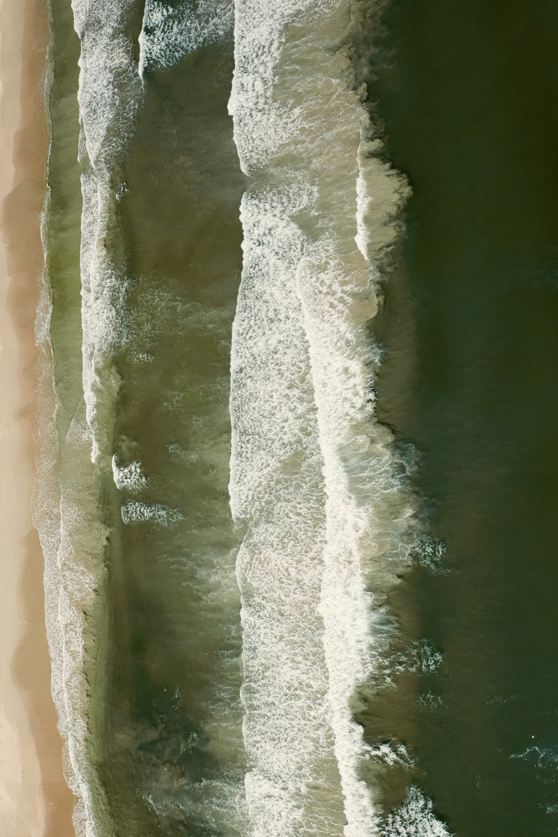 Aerial view of ocean waves with white foam rolling over dark green water next to a strip of sandy beach.