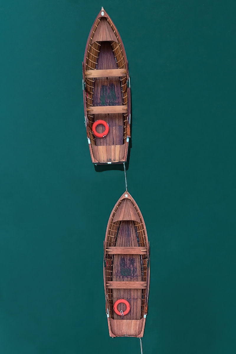 Two wooden rowboats tied together in a line over deep teal water, viewed from directly above. Each boat has a red life preserver.