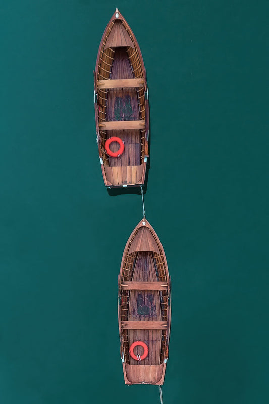 Two wooden rowboats tied together in a line over deep teal water, viewed from directly above. Each boat has a red life preserver.