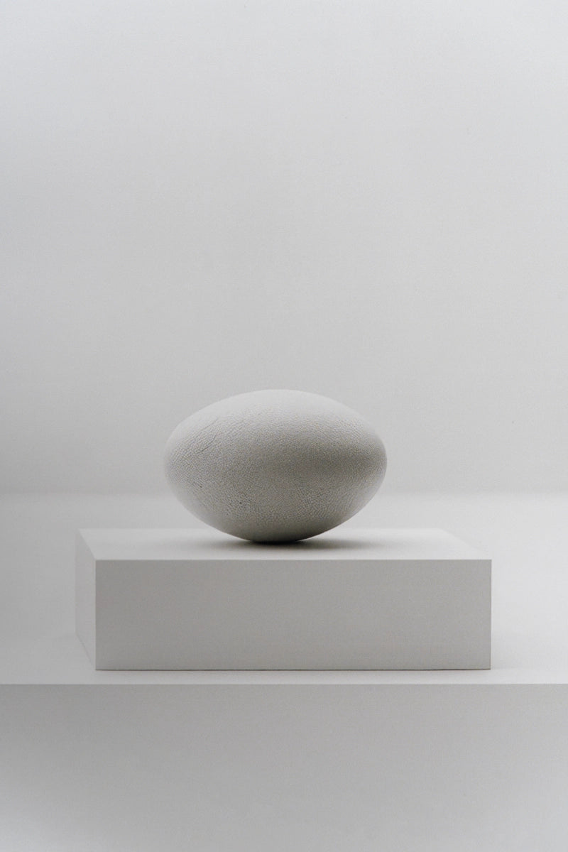 A minimalist light gray stone sculpture resembling a smooth egg, displayed on a matching rectangular pedestal against a clean, neutral white background.