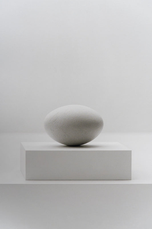 A minimalist light gray stone sculpture resembling a smooth egg, displayed on a matching rectangular pedestal against a clean, neutral white background.