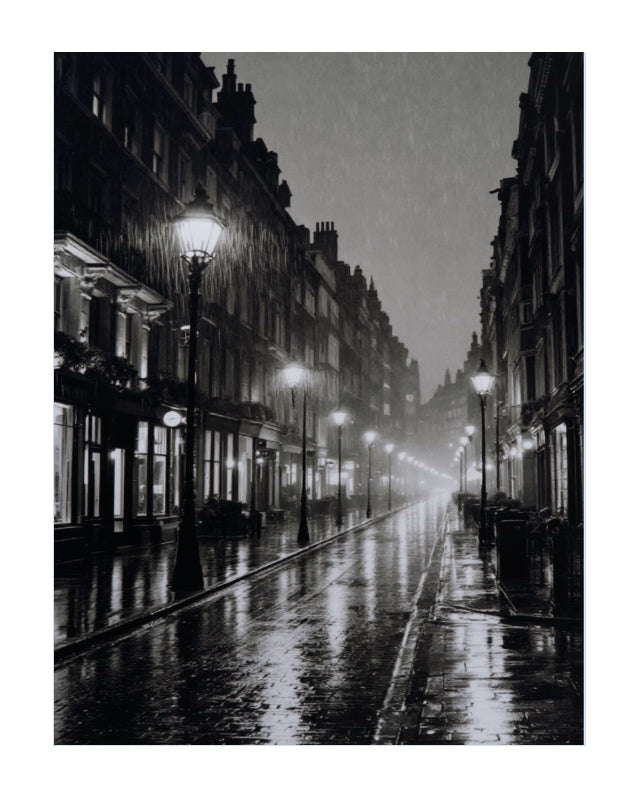 A black and white photograph of a wet city street lined with lit lampposts and tall buildings, presented as an art print with a white border.