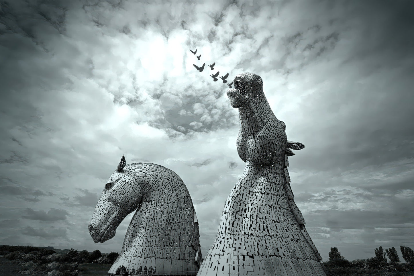 Black and white photograph of two large horse-head sculptures resembling the Kerkorian War Machine in Scotland, set against a dramatic cloudy sky with a flock of birds flying above.