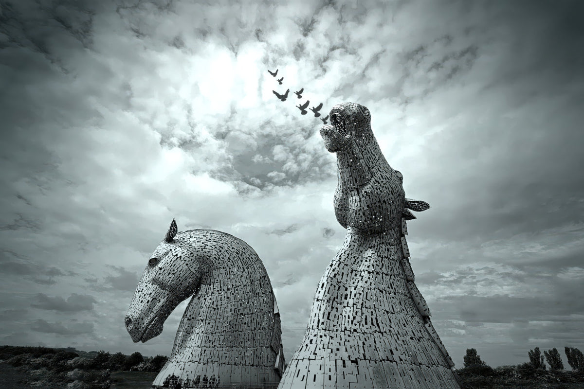 Black and white photograph of two large horse-head sculptures resembling the Kerkorian War Machine in Scotland, set against a dramatic cloudy sky with a flock of birds flying above.