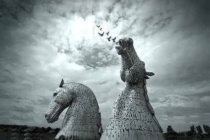 Black and white photograph of two large horse-head sculptures resembling the Kerkorian War Machine in Scotland, set against a dramatic cloudy sky with a flock of birds flying above.