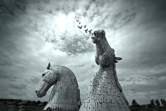 Black and white photograph of two large horse-head sculptures resembling the Kerkorian War Machine in Scotland, set against a dramatic cloudy sky with a flock of birds flying above.