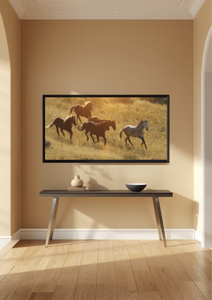 A framed wall art print showing five horses running through a sunlit grassy field, displayed above a dark console table in a warm beige room with wood flooring.