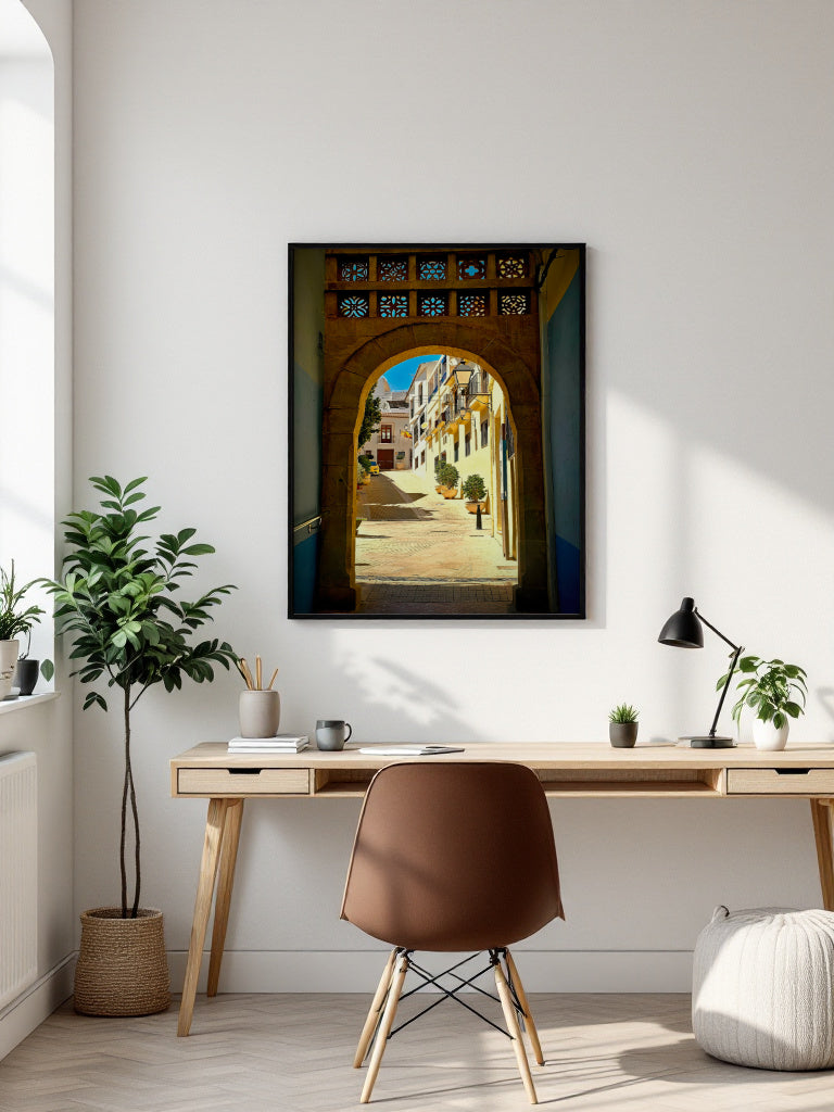 A framed vertical art print depicting a sunlit street scene through a stone archway, displayed on a white wall above a modern desk setup with a chair, lamp, and plants.