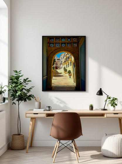 A framed vertical art print depicting a sunlit street scene through a stone archway, displayed on a white wall above a modern desk setup with a chair, lamp, and plants.