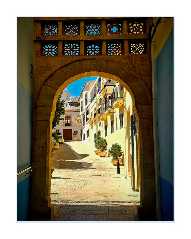 A scenic art print depicting a view from under a stone archway looking down a sunlit cobblestone street lined with yellow buildings and potted trees.