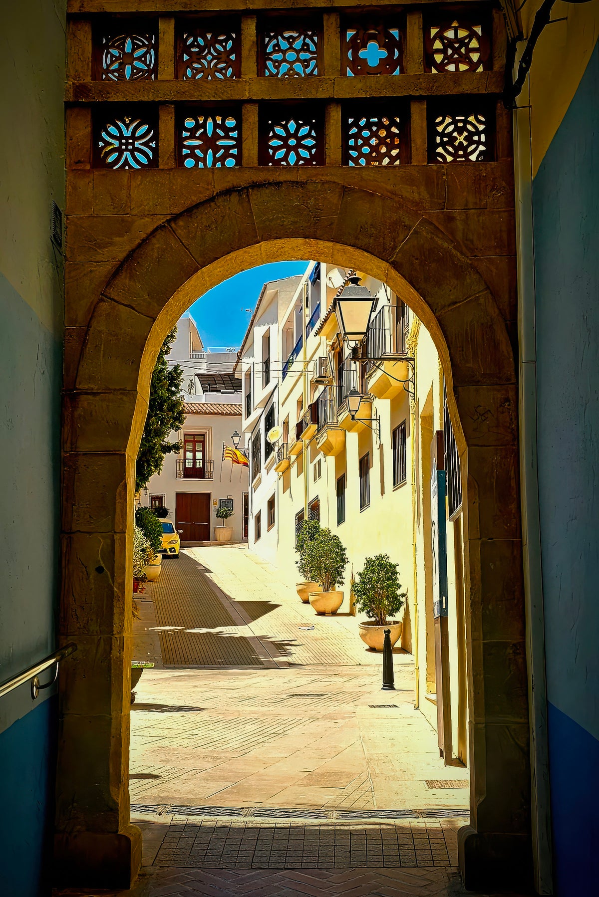 A stone archway frames a sunlit narrow street with yellow buildings, blue sky, and potted plants. The arch has decorative blue and orange geometric cutout panels at the top.