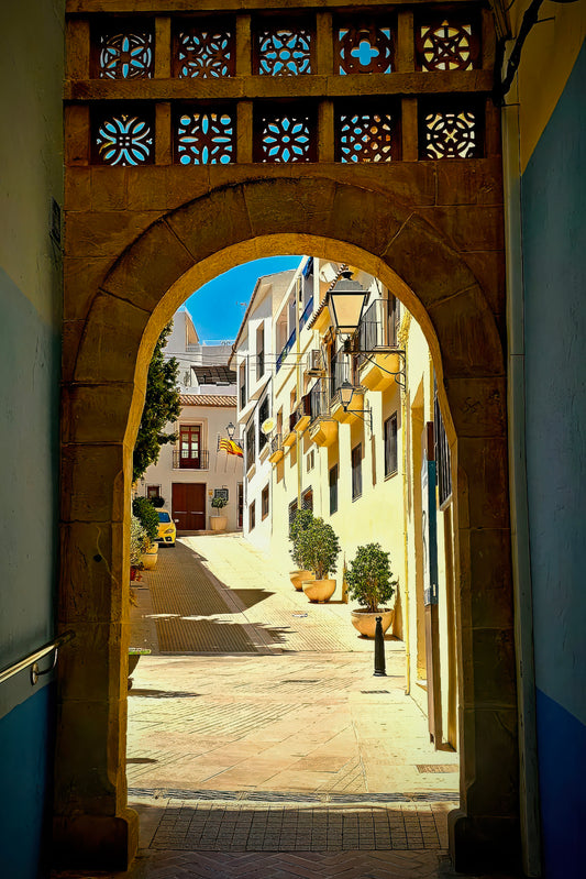 A stone archway frames a sunlit narrow street with yellow buildings, blue sky, and potted plants. The arch has decorative blue and orange geometric cutout panels at the top.