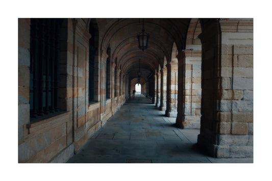 A scenic photograph of a stone colonnade with arches and columns, featuring a tiled floor leading to a bright opening in the distance, presented with a white border.